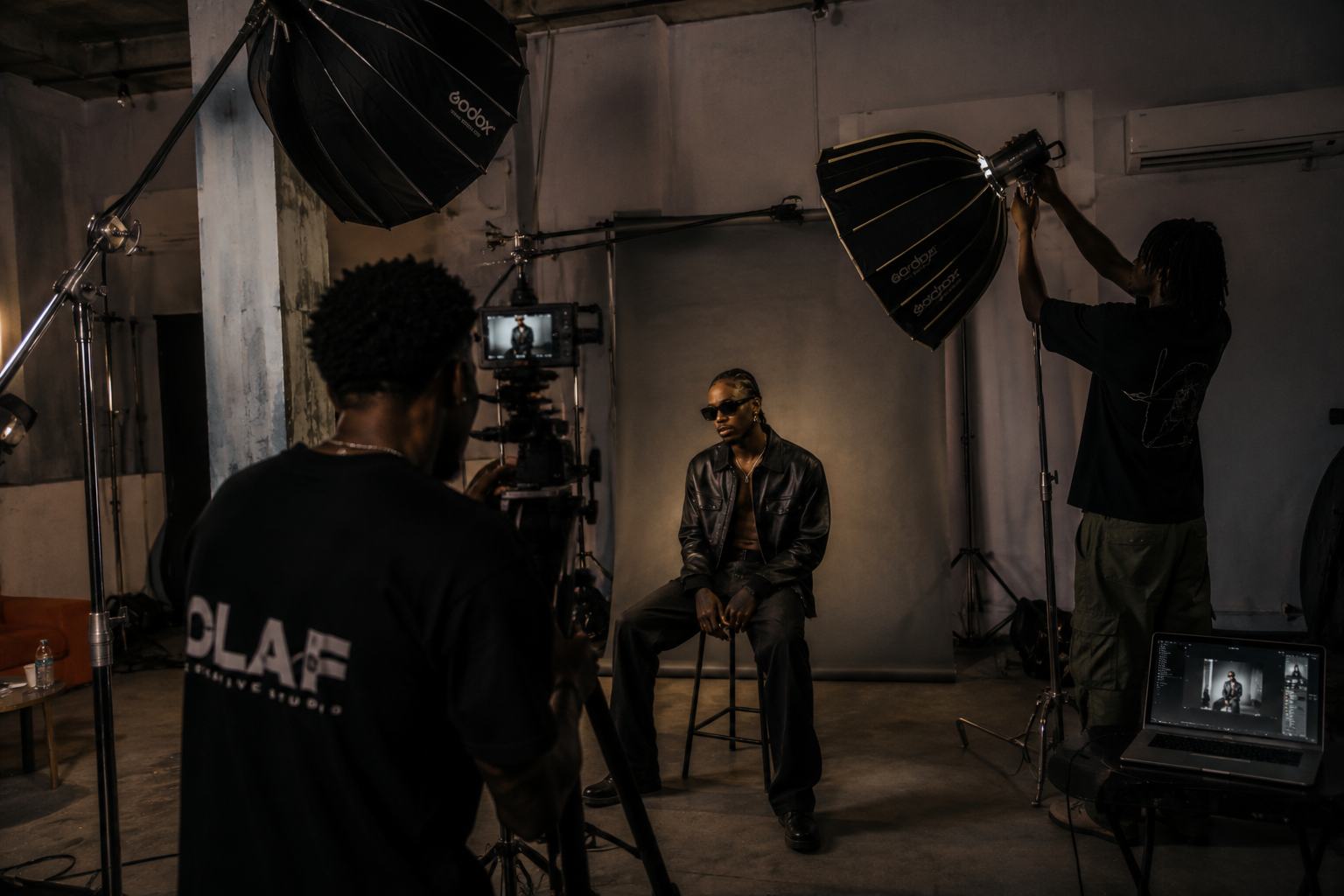 An OLAF Studio crew lighting a seated subject on a grey backdrop — two softboxes, a camera on sticks, a tethered laptop.